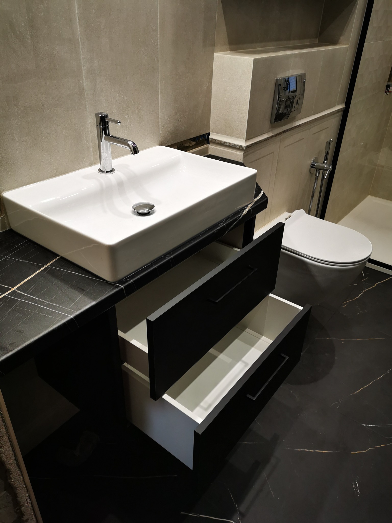 Nowoczesna łazienka z czarnym blatem imitującym marmur, białą umywalką nablatową i otwartymi szufladami. Minimalistyczny design z widoczną toaletą w tle.
