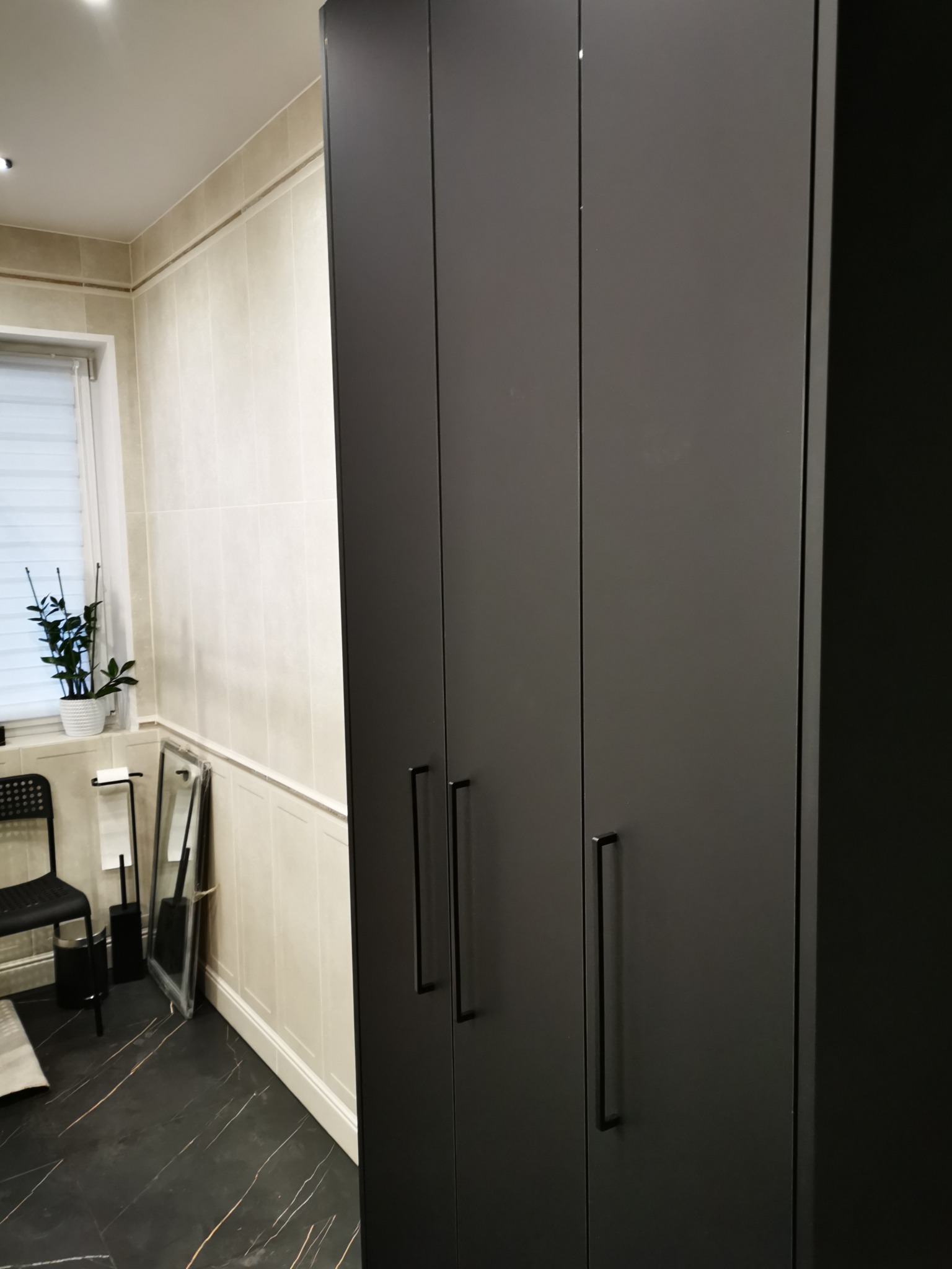 Ciemnoszara szafa trzydrzwiowa z podłużnymi uchwytami w łazience z czarną marmurową podłogą i jasnymi płytkami ściennymi. Widoczny fragment rośliny i akcesoriów łazienkowych.