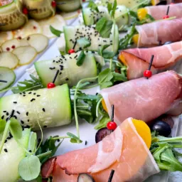 Elegancko zaaranżowane przekąski na białym talerzu: ruloniki z og&oacute;rka z farszem, plasterki og&oacute;rka z serem i czarnym sezamem oraz szynka parmeńska zawijana na kawałkach owoc&oacute;w, wszystko udekorowane...