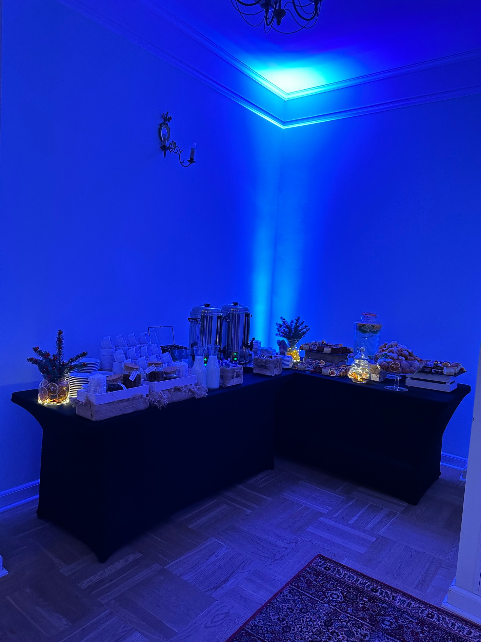 Stół cateringowy z przekąskami, napojami i dekoracjami świątecznymi podświetlony niebieskim światłem, widoczny dywan na podłodze.