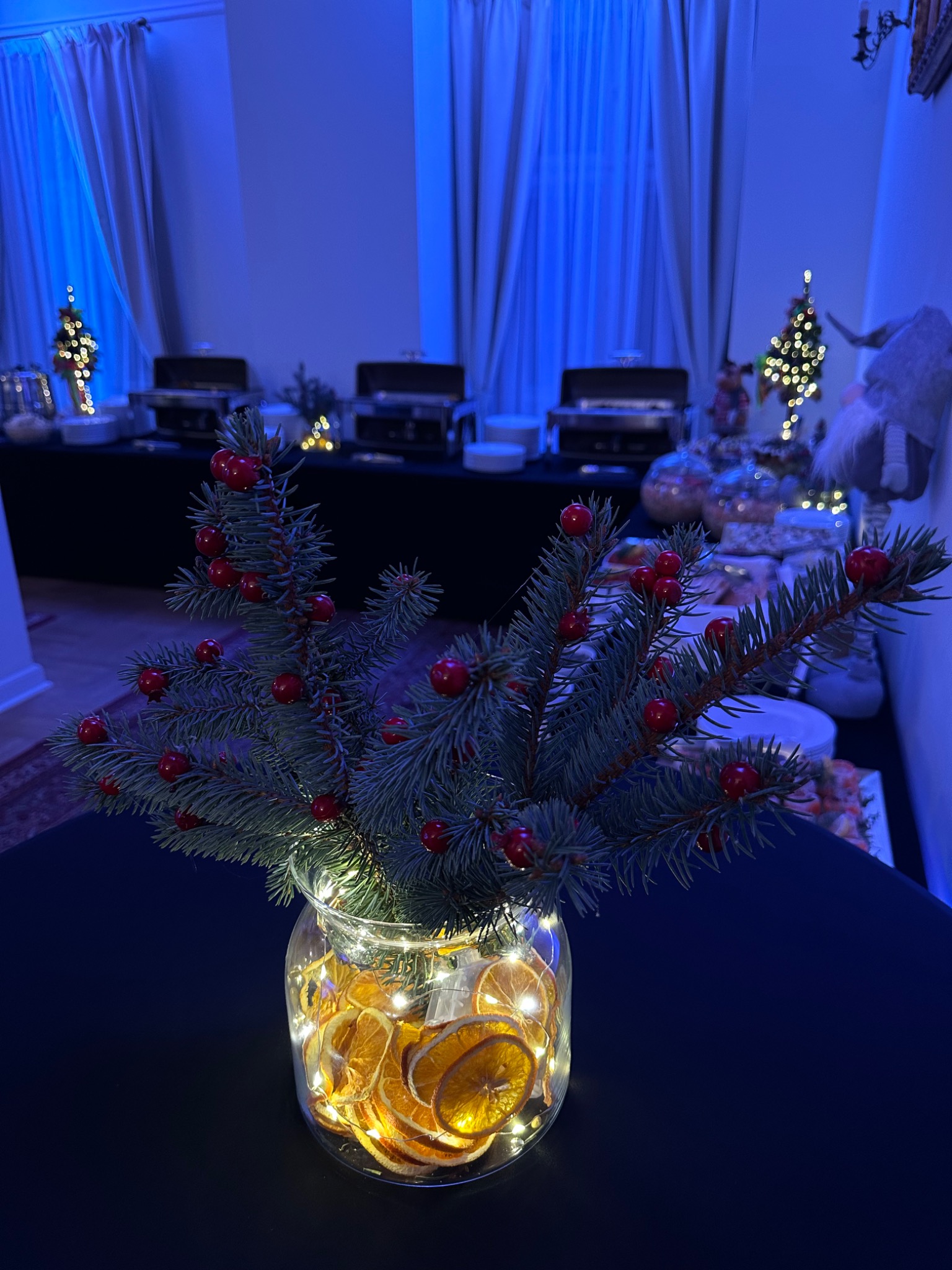 Świąteczna dekoracja stołu: gałązka świerku z czerwonymi owocami w szklanym wazonie z suszonymi pomarańczami i lampkami, bufet z podgrzewaczami potraw w tle, niebieskie oświetlenie.