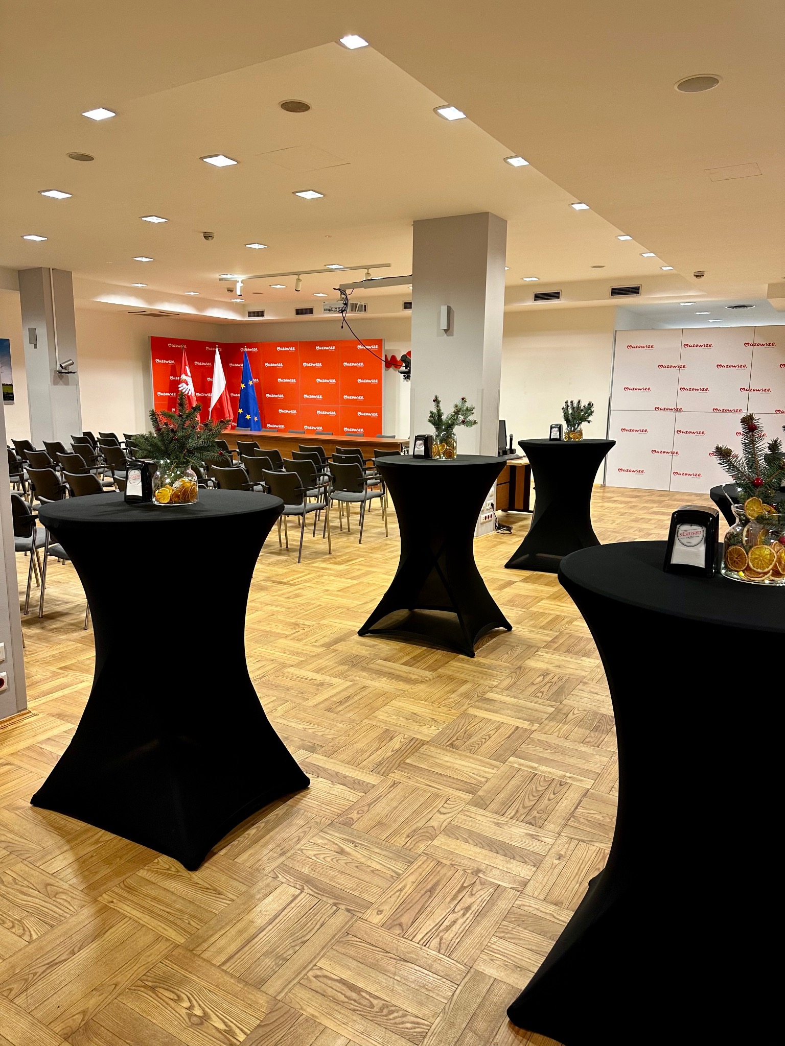 Sala konferencyjna przygotowana na wydarzenie z ustawionymi stolikami koktajlowymi z czarnymi obrusami, dekoracjami z gałązek i suszonych pomarańczy, w tle krzesła i flaga Polski, Unii Europejskiej...