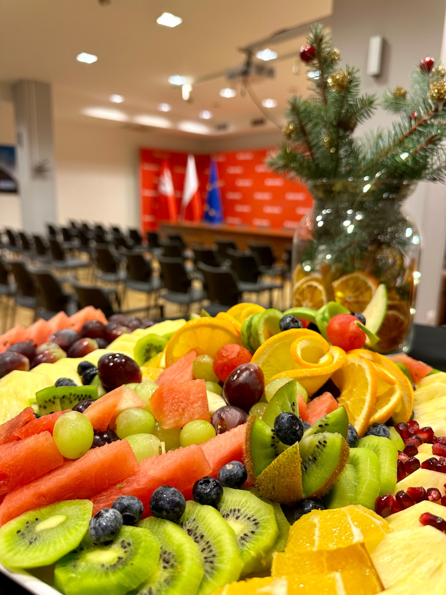 Bogata kompozycja owocowa z arbuzem, winogronami, kiwi, pomarańczami i borówkami na tle sali konferencyjnej z flagami i świąteczną dekoracją w szklanym wazonie.