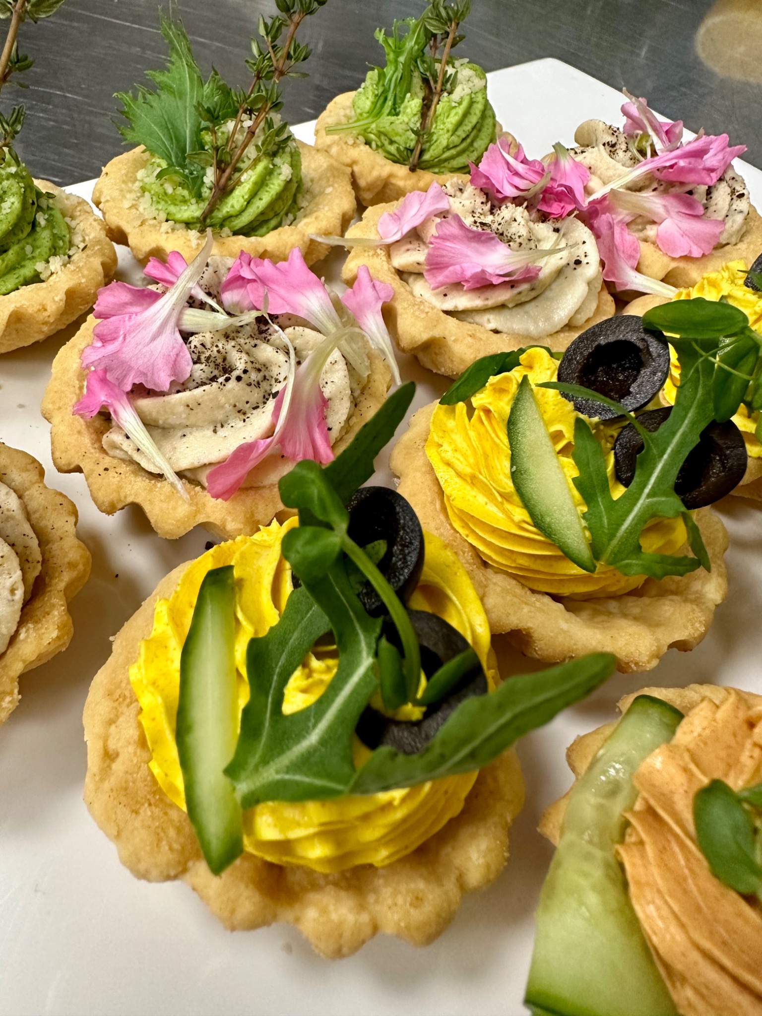 Elegancka kompozycja różnorodnych tartaletek z kremowymi nadzieniami w odcieniach zieleni, żółci i beżu, ozdobionych świeżymi ziołami, plastrami ogórka, oliwkami i delikatnymi płatkami różowych...