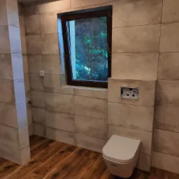 Nowoczesna łazienka z beżowymi, podłużnymi płytkami na ścianach, drewnopodobną podłogą i oknem z widokiem na zieleń, częściowo zamontowana toaleta podtynkowa.