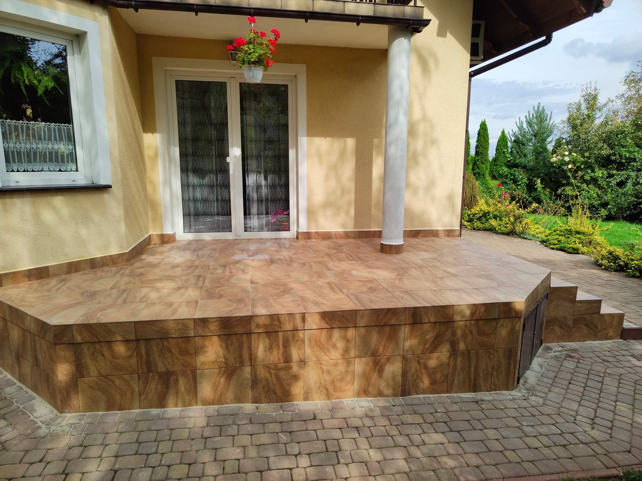 Wykafelkowany taras przed domem z kremową elewacją, widoczna kolumna, okno z firanką i drzwi balkonowe z kwiatami w doniczce wiszącej.