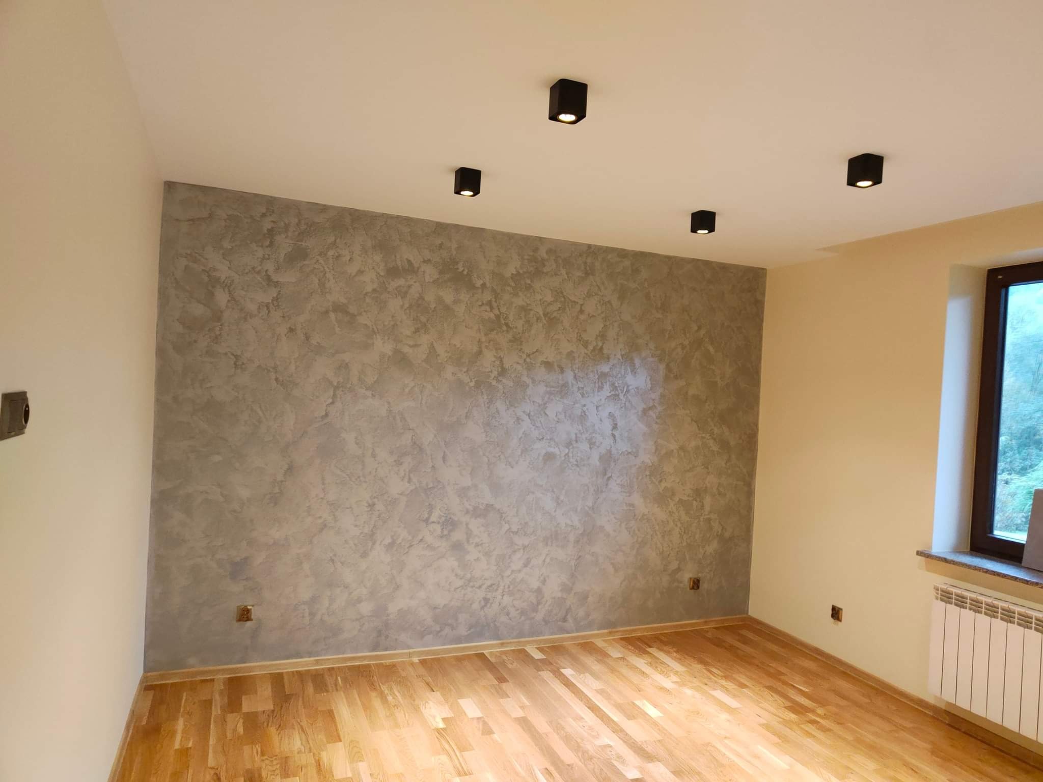 Pokój po remoncie: ściana z efektem dekoracyjnym w odcieniach szarości, drewniana podłoga, czarne, kwadratowe lampy sufitowe, biały grzejnik pod oknem.