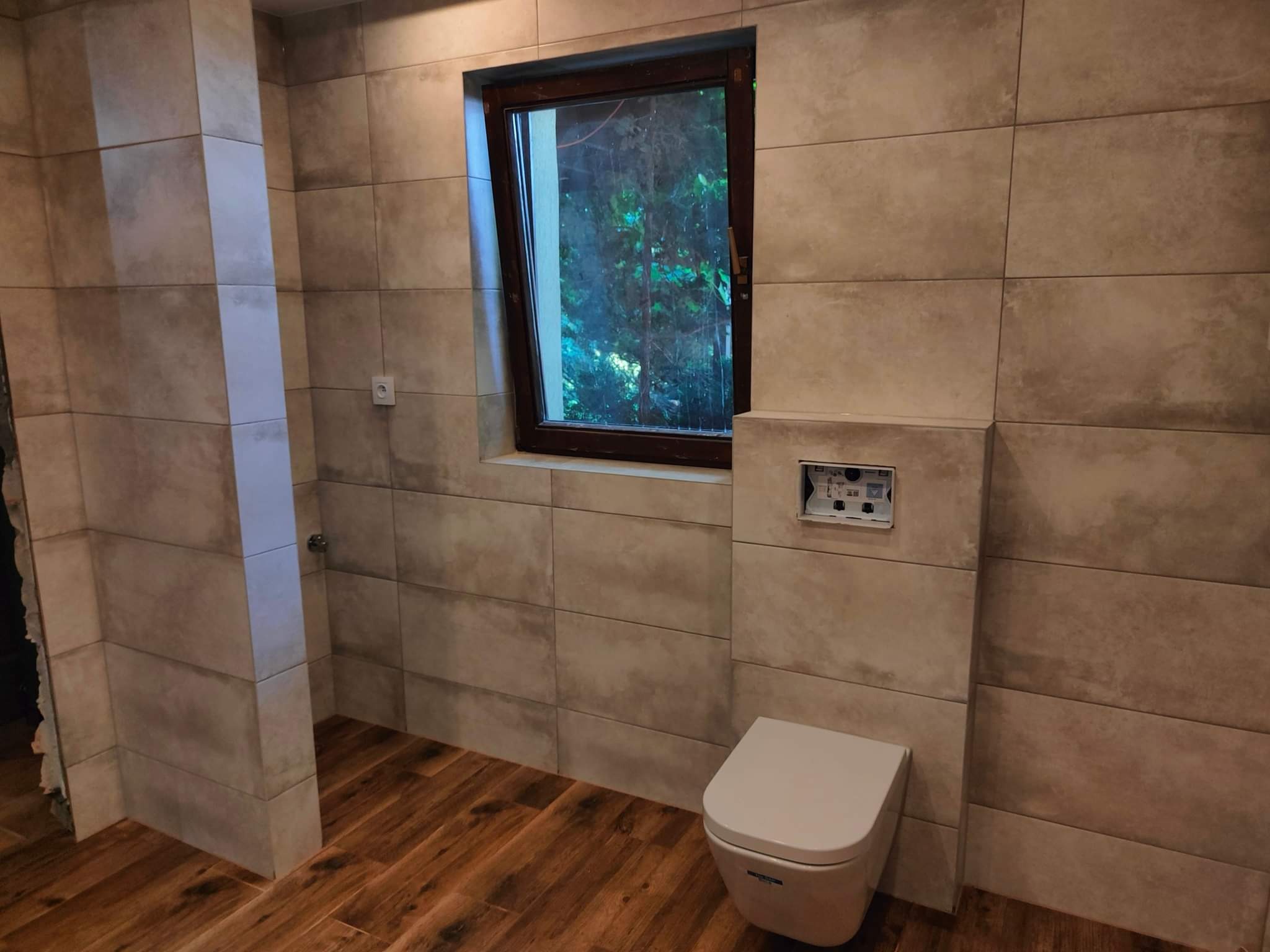 Nowoczesna łazienka z beżowymi, podłużnymi płytkami na ścianach, drewnopodobną podłogą i oknem z widokiem na zieleń, częściowo zamontowana toaleta podtynkowa.