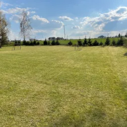 Rozległy trawnik z widocznymi śladami koszenia, bramki do piłki nożnej, trampolina i rząd iglaków w tle, błękitne niebo z obłokami.