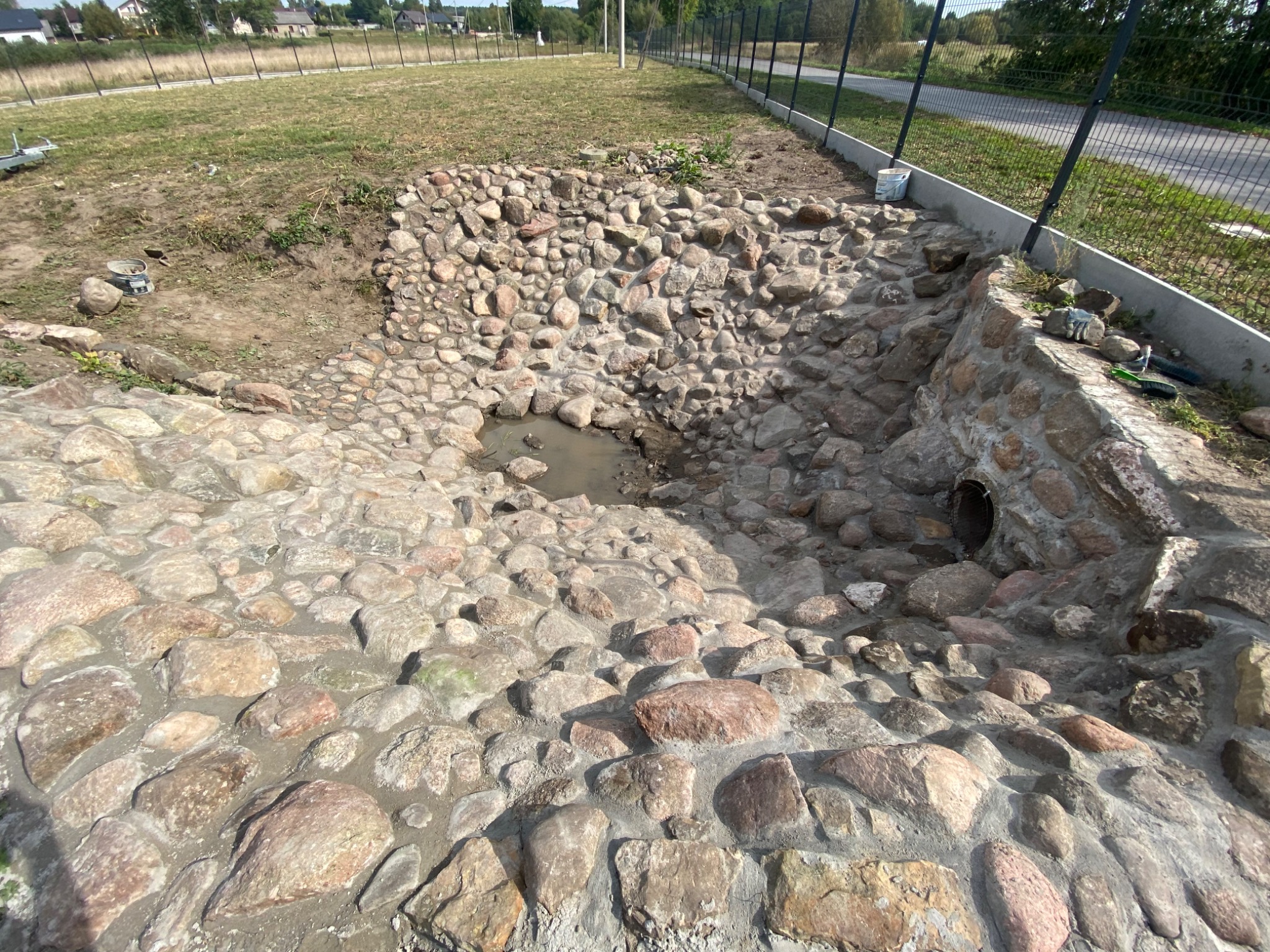 Budowa skalniaka z kamieni polnych na trawniku, widoczne narzędzia i wiadro z zaprawą, fragment ogrodzenia w tle.