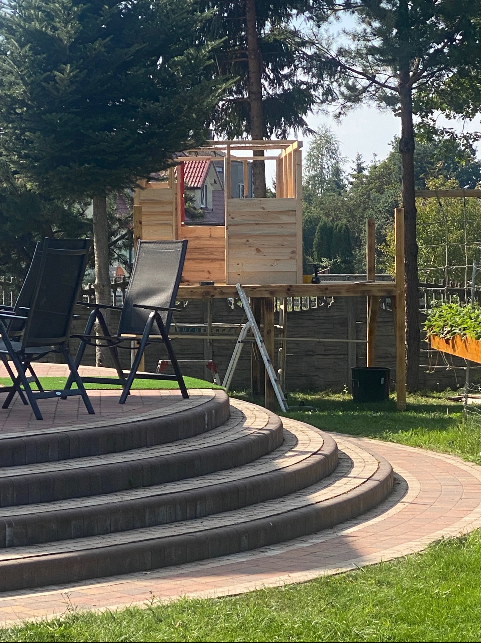 Drewniana konstrukcja domku na drzewie w trakcie budowy, widoczna z tarasu z krzesłami ogrodowymi, z elementami rusztowania i narzędziami.