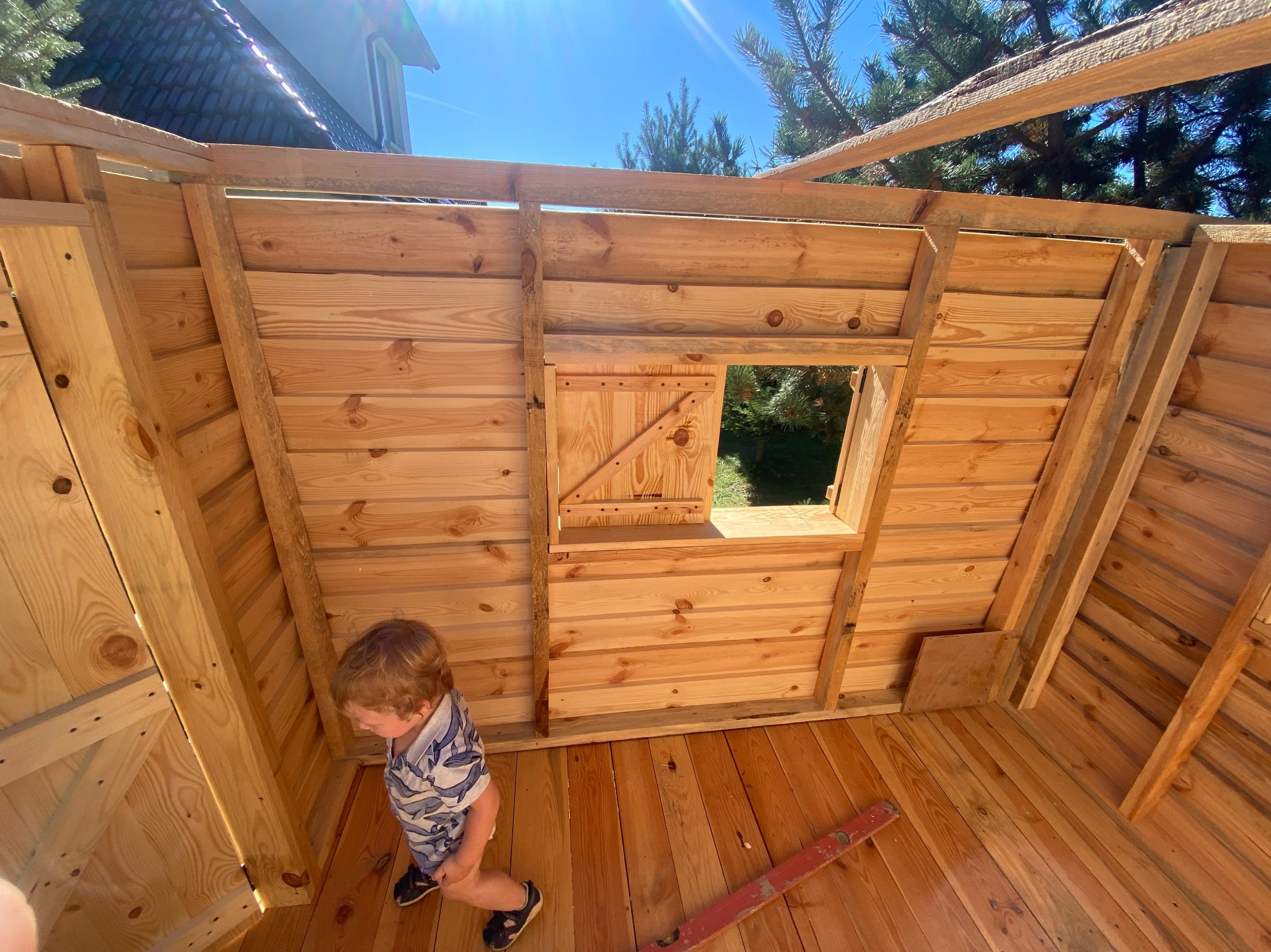 Wnętrze drewnianej konstrukcji z widocznym oknem z okiennicą i fragmentem dachu, w środku stoi małe dziecko, a na podłodze leży poziomica.