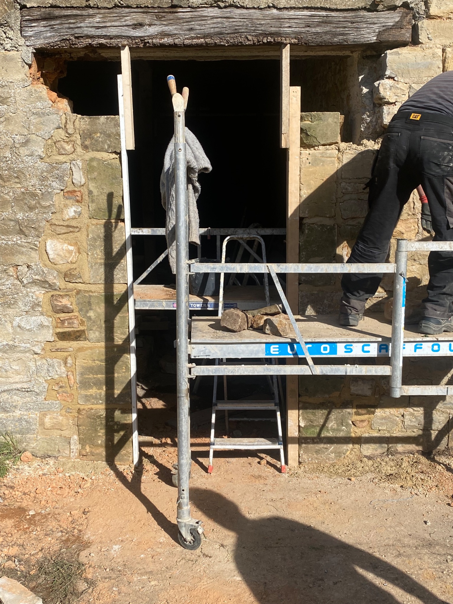 Renowacja kamiennego muru z użyciem rusztowania, widoczny fragment ściany z ościeżnicą, narzędzia i ubranie robocze