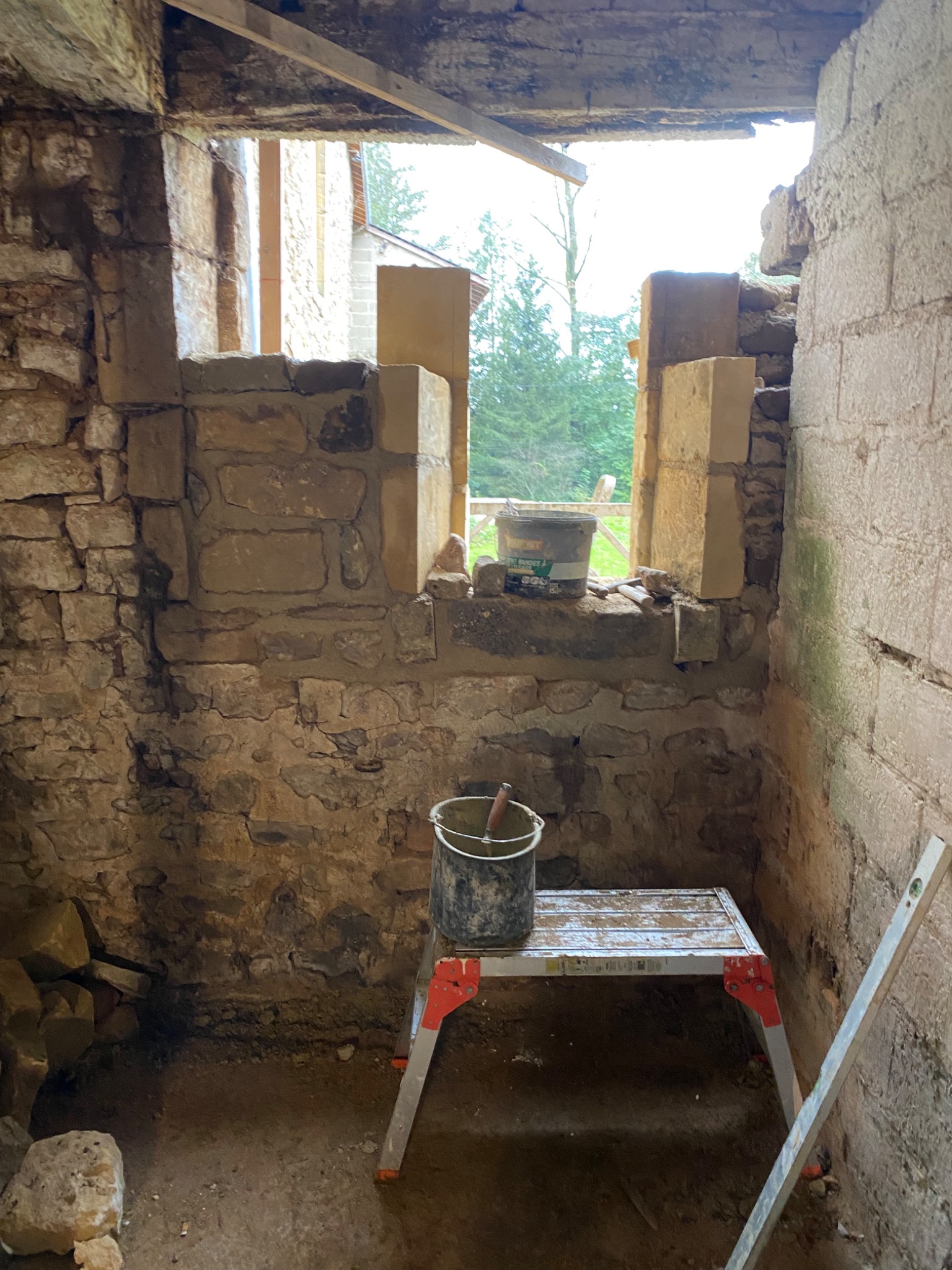 Wnętrze kamiennego budynku w trakcie renowacji okna, widoczna drabina, poziomica i wiadro z zaprawą.