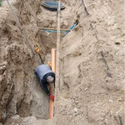 Pracownik w wykopie, w czerwonych rękawicach, łączy pomarańczową rurę kanalizacyjną; widoczne zabezpieczenia wykopu, inne rury i ubijarka w tle.