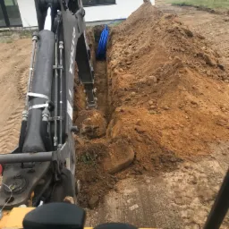 Widok z kabiny operatora koparki Volvo na wykop z niebieskimi rurami, prawdopodobnie pod przyłącze wodno-kanalizacyjne, na tle domu jednorodzinnego i zielonego wzgórza z samochodem.