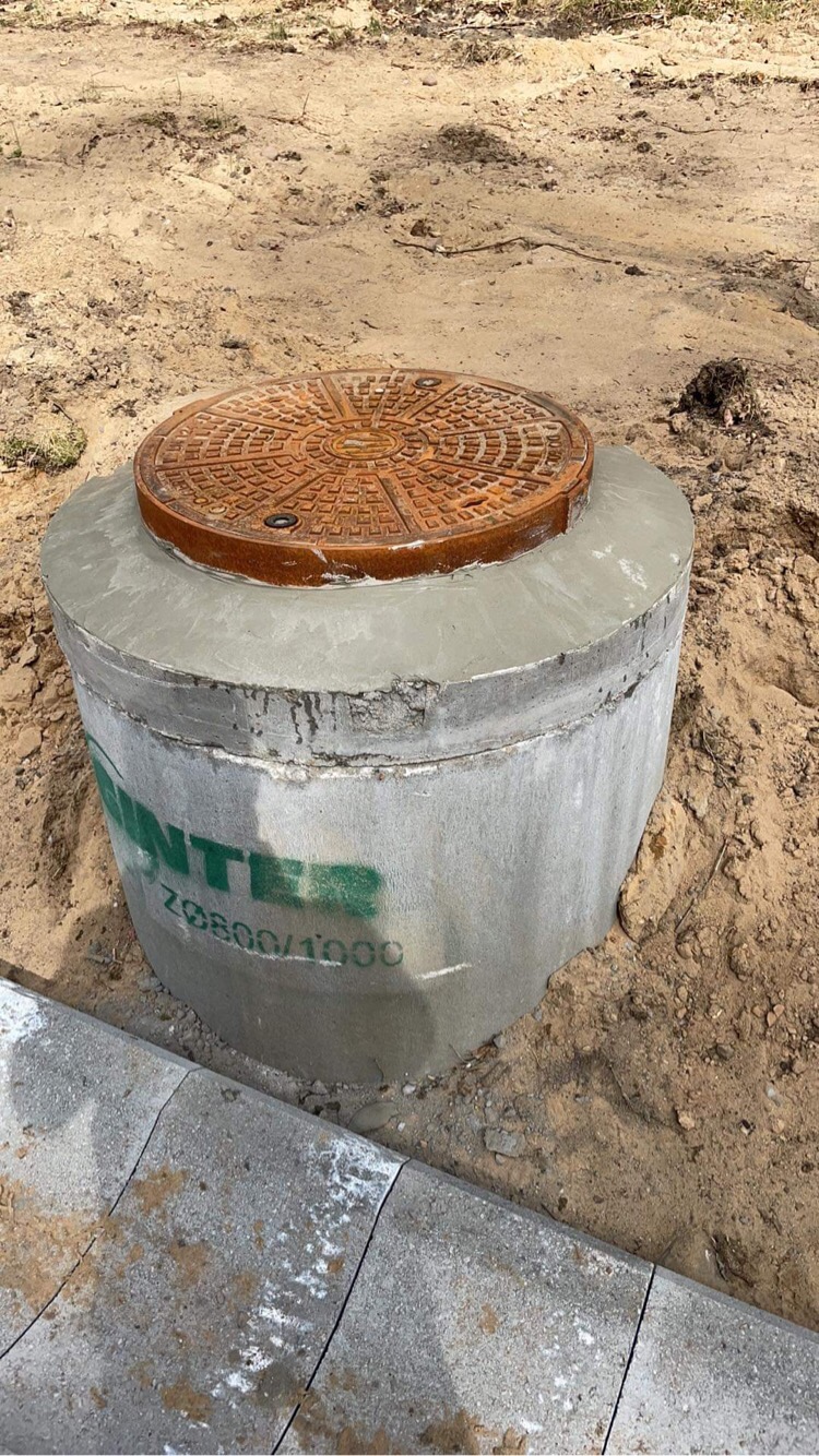 Betonowy zbiornik z żeliwną pokrywą włazu, umieszczony w piaszczystym gruncie obok ułożonej kostki brukowej.