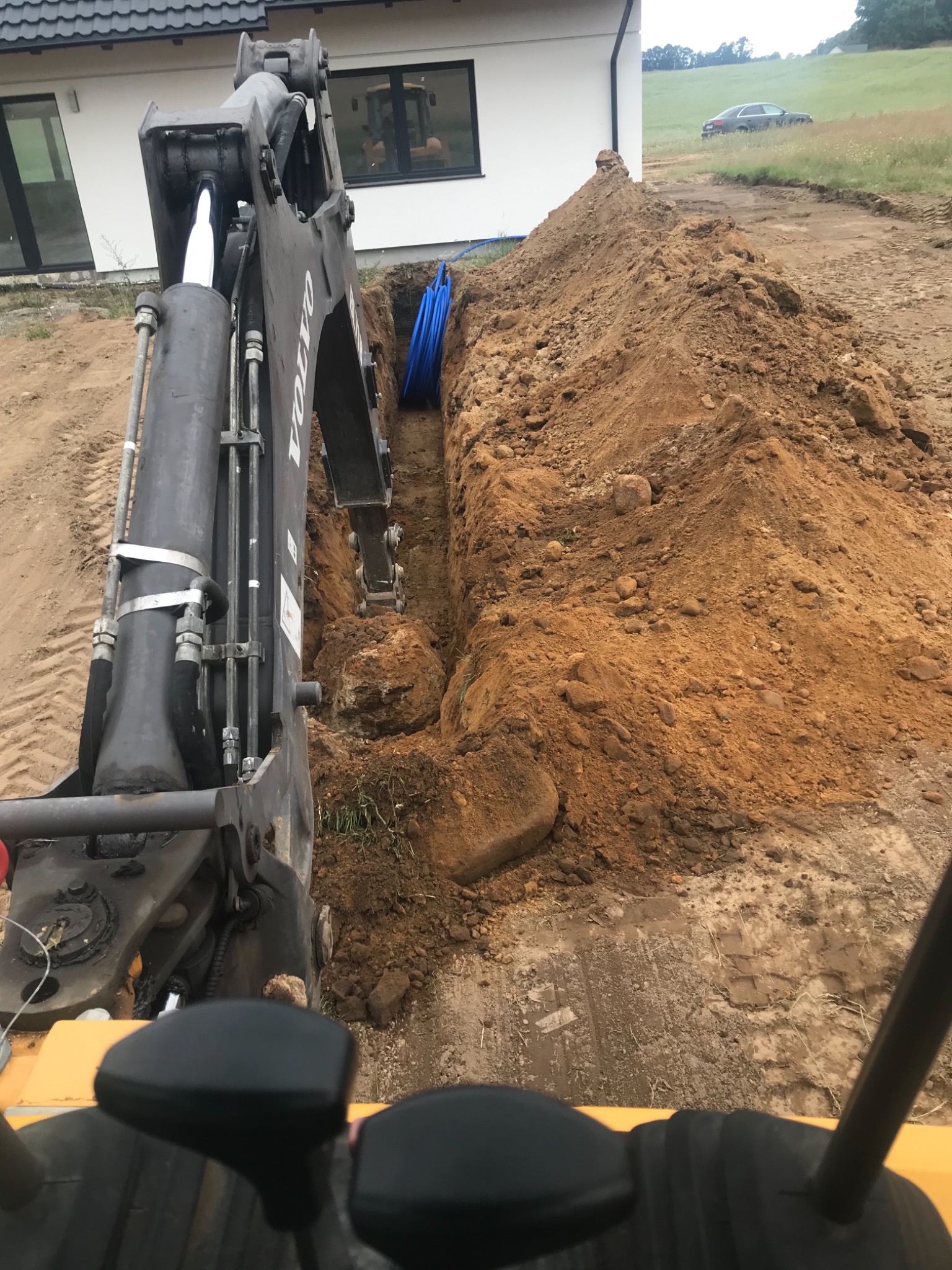 Widok z kabiny operatora koparki Volvo na wykop z niebieskimi rurami, prawdopodobnie pod przyłącze wodno-kanalizacyjne, na tle domu jednorodzinnego i zielonego wzgórza z samochodem.