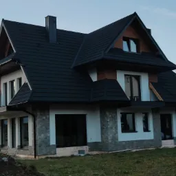 Nowoczesny dom jednorodzinny z czarnym dachem i elewacją w odcieniach bieli i szarości, widok z zewnątrz na częściowo wykończoną elewację z widoczną izolacją u podstawy.