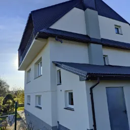 Nowoczesny, dwupiętrowy dom z balkonem i ciemnym dachem w Białym Dunajcu po świeżym tynkowaniu, z widocznym wiaderkiem farby i resztkami materiałów budowlanych przy ścianie.