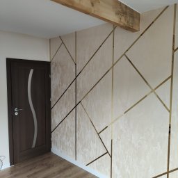 Jamrozowicz - Docieplenia & Wykończenia - Eleganckie wykończenie wnętrza: ściana z beżowym tynkiem strukturalnym, zdobiona złotymi listwami tworzącymi geometryczny wzór; widoczny drewniany belka stropowa z dekoracyjnymi wzorami i fragment...