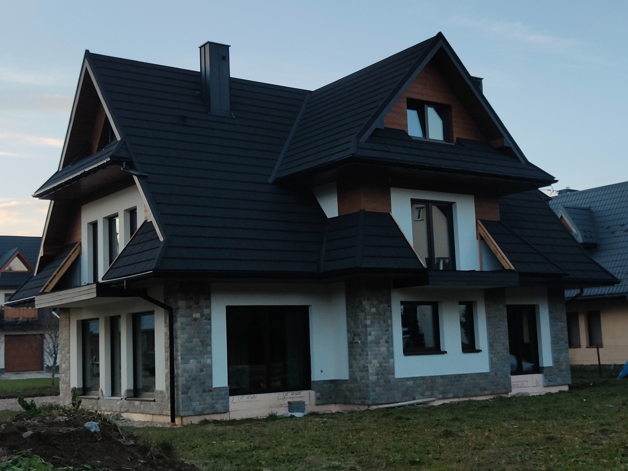 Nowoczesny dom jednorodzinny z czarnym dachem i elewacją w odcieniach bieli i szarości, widok z zewnątrz na częściowo wykończoną elewację z widoczną izolacją u podstawy.