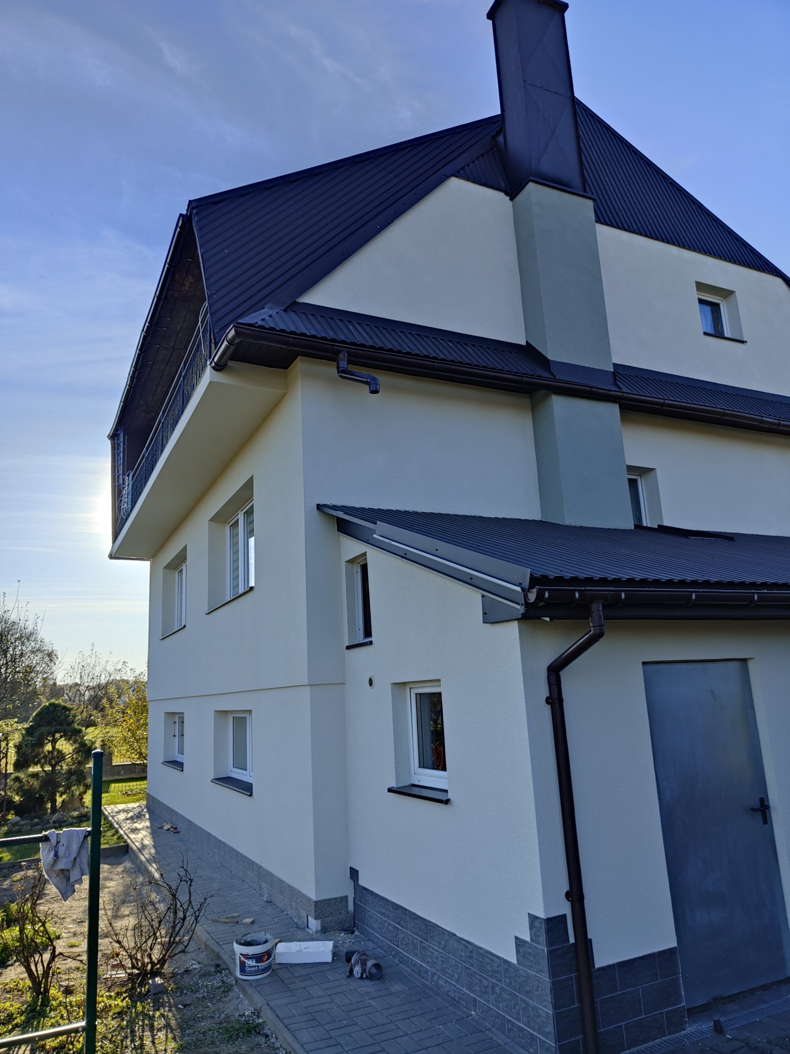 Nowoczesny, dwupiętrowy dom z balkonem i ciemnym dachem w Białym Dunajcu po świeżym tynkowaniu, z widocznym wiaderkiem farby i resztkami materiałów budowlanych przy ścianie.