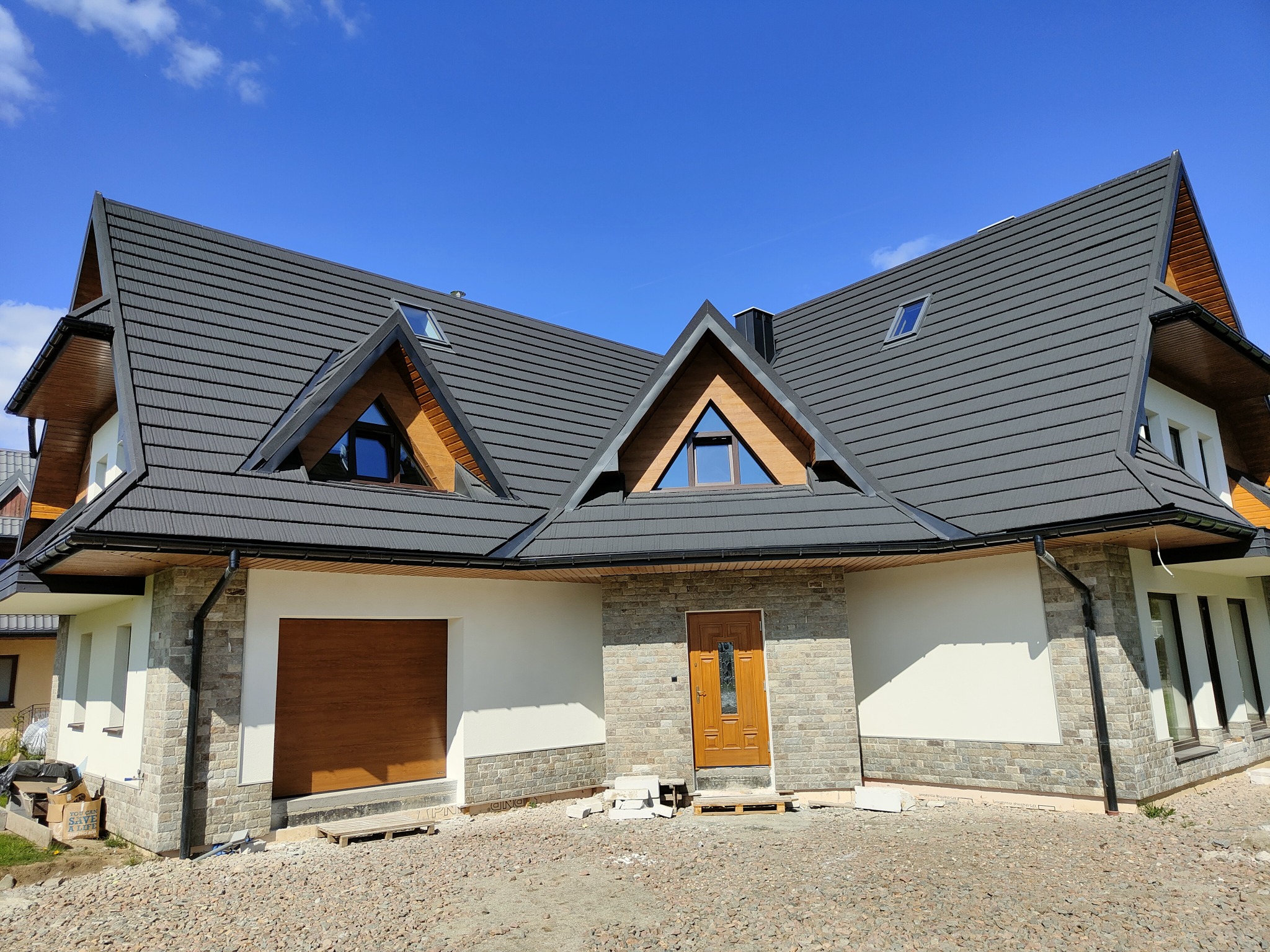 Nowoczesny dom z szarą dachówką i elementami kamiennymi na elewacji. Widoczne okna dachowe i drewniana stolarka okienna. Teren wokół domu w trakcie prac wykończeniowych.
