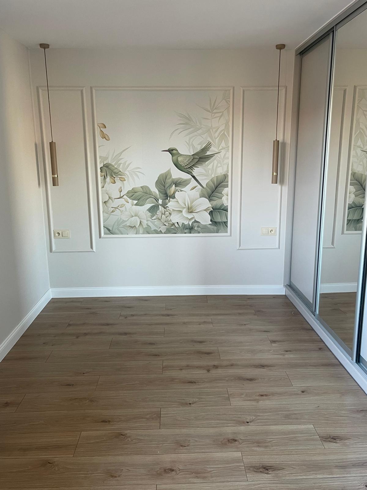 Jasne panele podłogowe w pokoju z dekoracyjną ścianą z motywem ptaka i kwiatów, oświetloną przez wiszące lampy. Minimalistyczny design z szafą z lustrzanymi drzwiami.