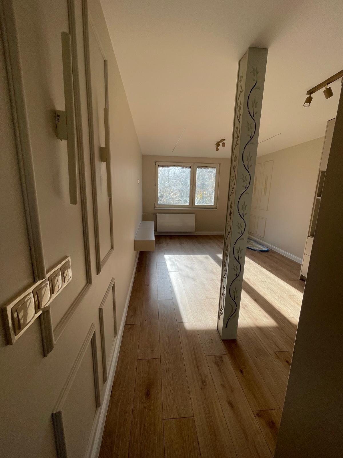 Jasne wnętrze po remoncie: drewnopodobne panele, dekoracyjna ściana z listwami, kolumna z ornamentem, okno z widokiem na drzewa. Świeżo i nowocześnie.