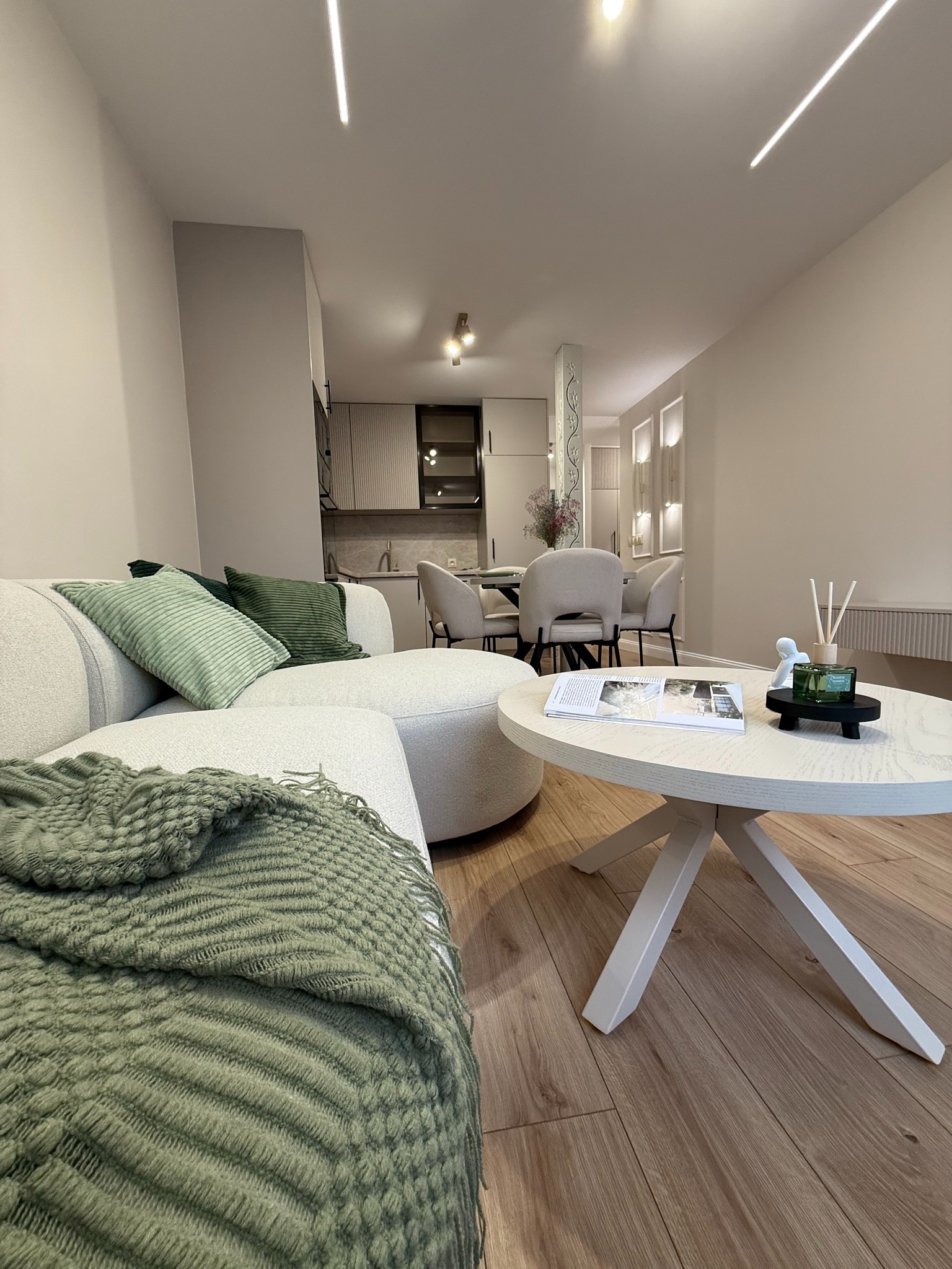 Jasne wnętrze: sofa z zielonymi poduszkami i pledem, okrągły stolik z książką i dekoracjami. W tle widoczna kuchnia i jadalnia w stonowanych barwach.