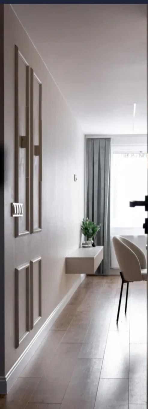 Nowoczesny przedpokój w jasnych barwach: drewnopodobna podłoga, minimalistyczna konsola ścienna z rośliną, dekoracyjne panele ścienne i krzesło przy oknie.