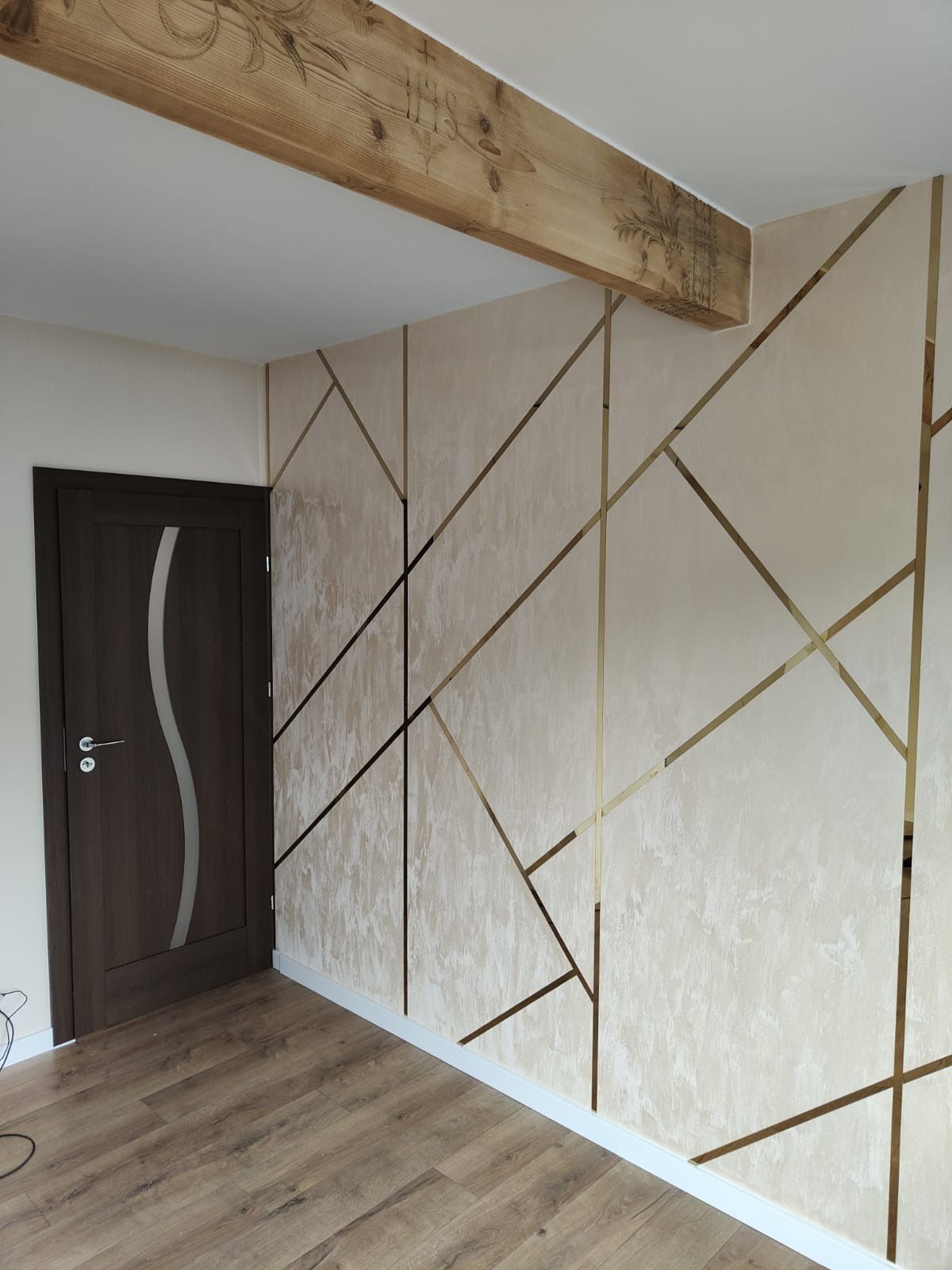 Eleganckie wykończenie wnętrza: ściana z beżowym tynkiem strukturalnym, zdobiona złotymi listwami tworzącymi geometryczny wzór; widoczny drewniany belka stropowa z dekoracyjnymi wzorami i fragment...