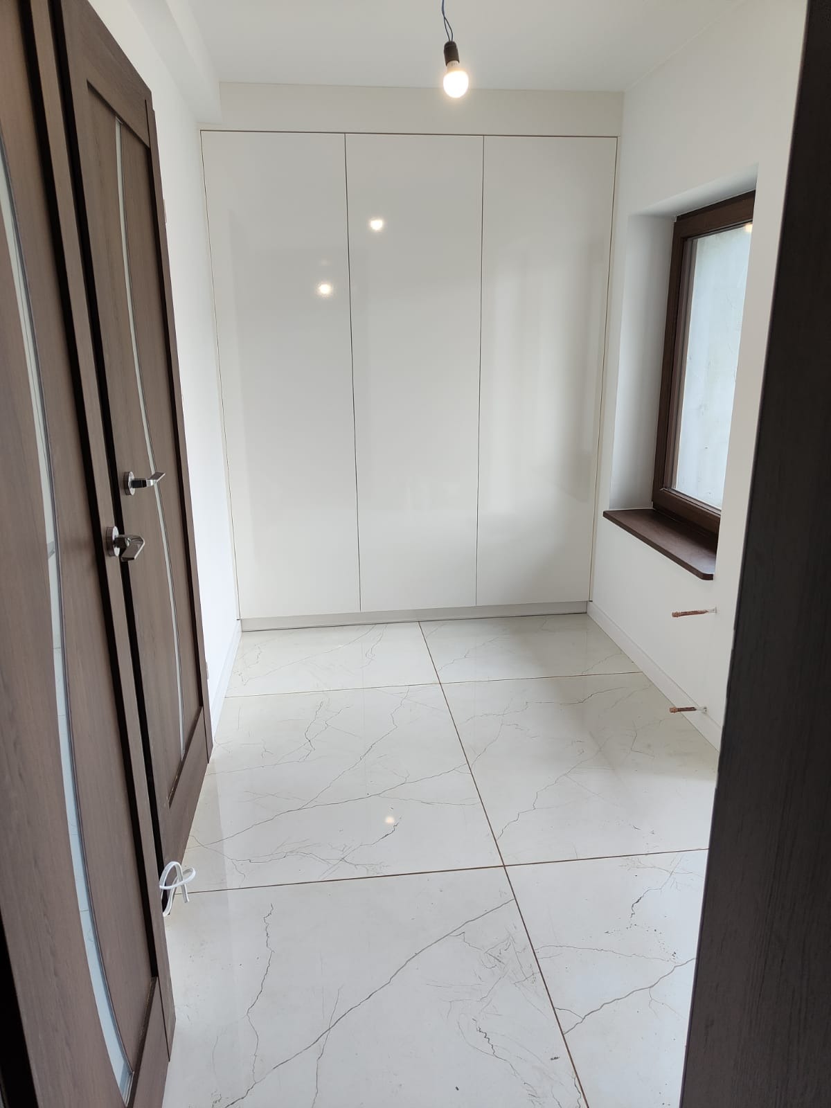 Nowoczesne wnętrze z białymi, lakierowanymi szafami w zabudowie, marmurową podłogą z szarymi żyłami, drewnianą stolarką okienną i drzwiami z ciemnego drewna.