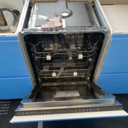 Dawid Szpejenkowski - Wnętrze otwartej zmywarki do naczyń z widocznymi koszami, dyszami natryskowymi i panelem sterowania, zamontowanej między niebieskimi szafkami kuchennymi, widok z góry.