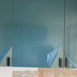 Dawid Szpejenkowski - Niebieskie szafki kuchenne z czarnymi uchwytami, częściowo zabezpieczone folią ochronną, zamontowane nad surową ścianą w trakcie prac wykończeniowych.