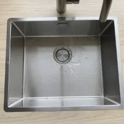 Nowa, prostokątna, stalowa zlewozmywak z baterią, zamontowana na blacie o strukturze jasnego drewna, widoczne krople wody w zlewie.