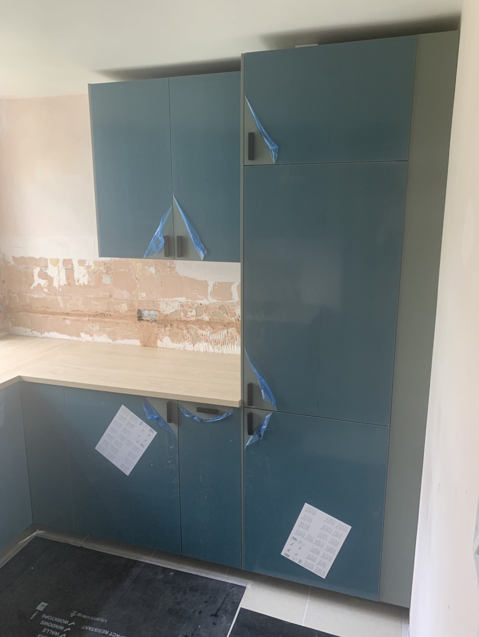 Montaż niebieskich szafek kuchennych z jasnym blatem, ściana w trakcie remontu z widoczną instalacją elektryczną, folia ochronna na uchwytach szafek.
