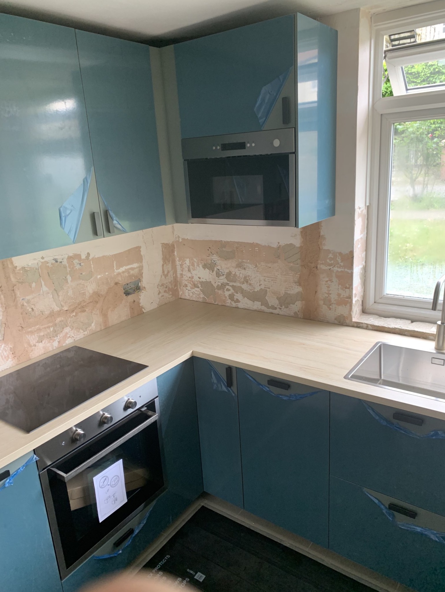Niedokończona kuchnia z niebieskimi szafkami zabezpieczonymi folią, surowe ściany przygotowywane do dalszych prac, blat z jasnego drewna, okno z widokiem na zieleń na zewnątrz.