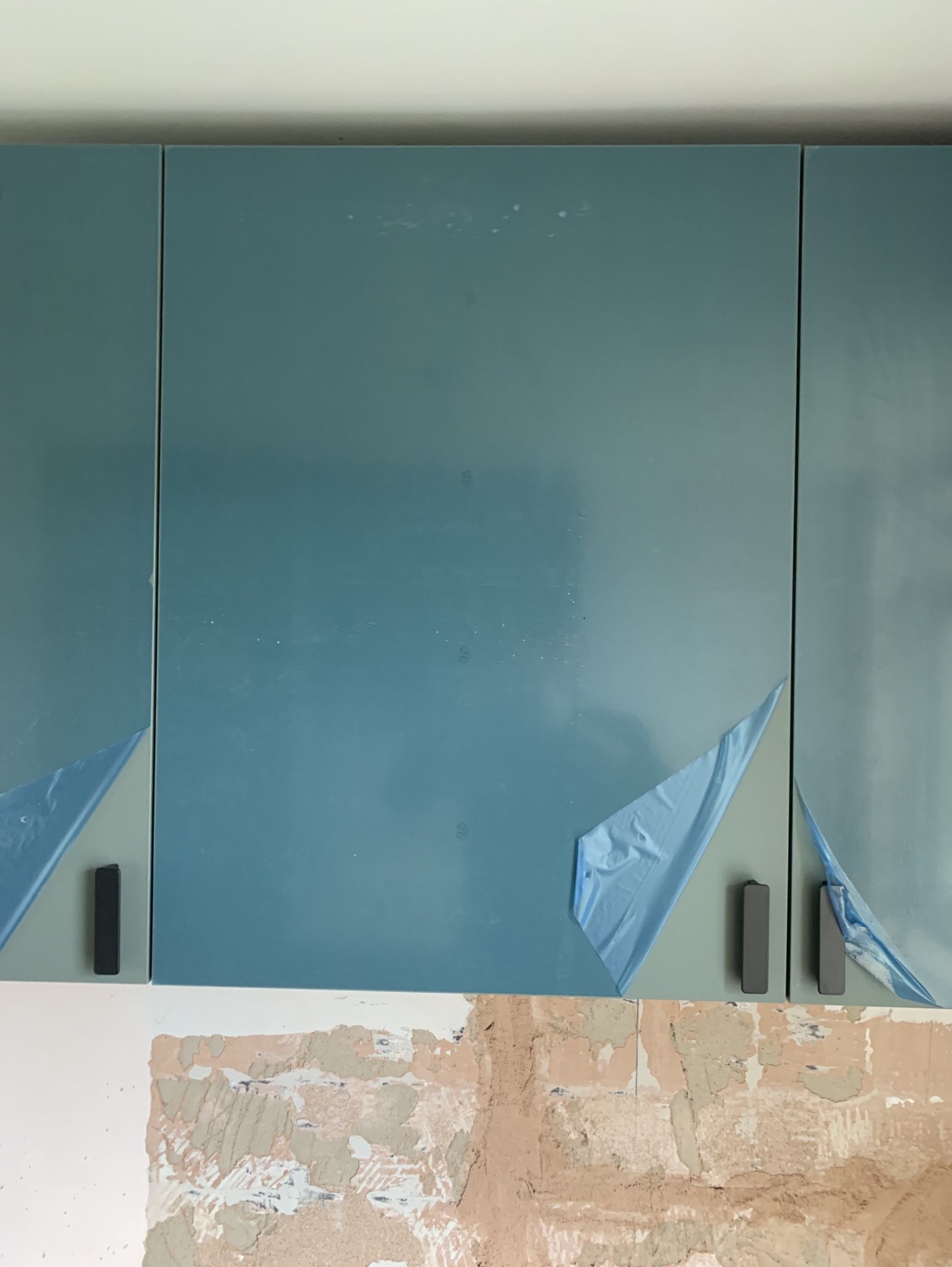 Niebieskie szafki kuchenne z czarnymi uchwytami, częściowo zabezpieczone folią ochronną, zamontowane nad surową ścianą w trakcie prac wykończeniowych.