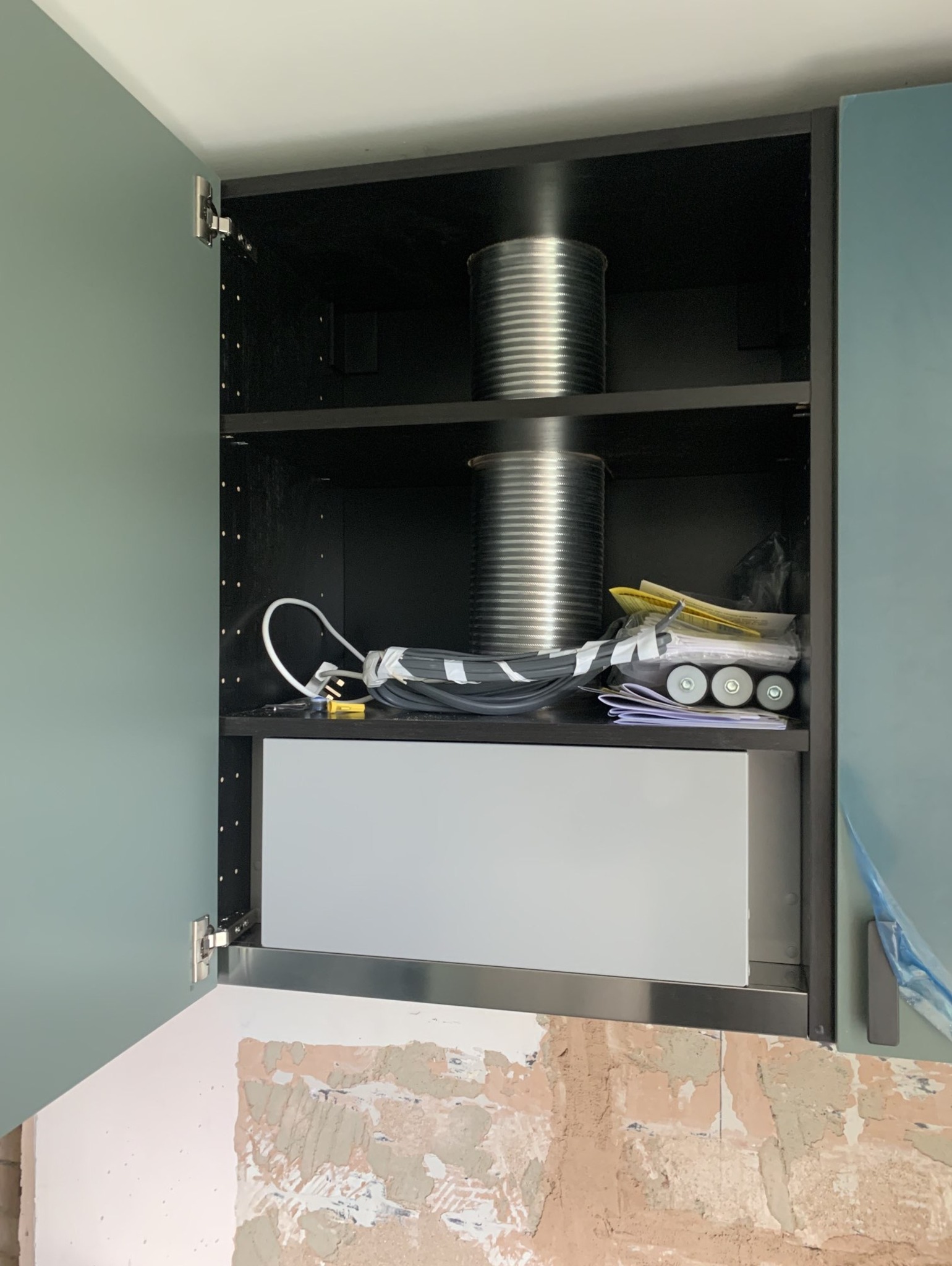 Wnętrze szafki kuchennej z otwartymi drzwiczkami, widoczne dwa metalowe przewody wentylacyjne, zwój kabli elektrycznych, dokumenty i trzy rolki, ściana za szafką w trakcie prac wykończeniowych.
