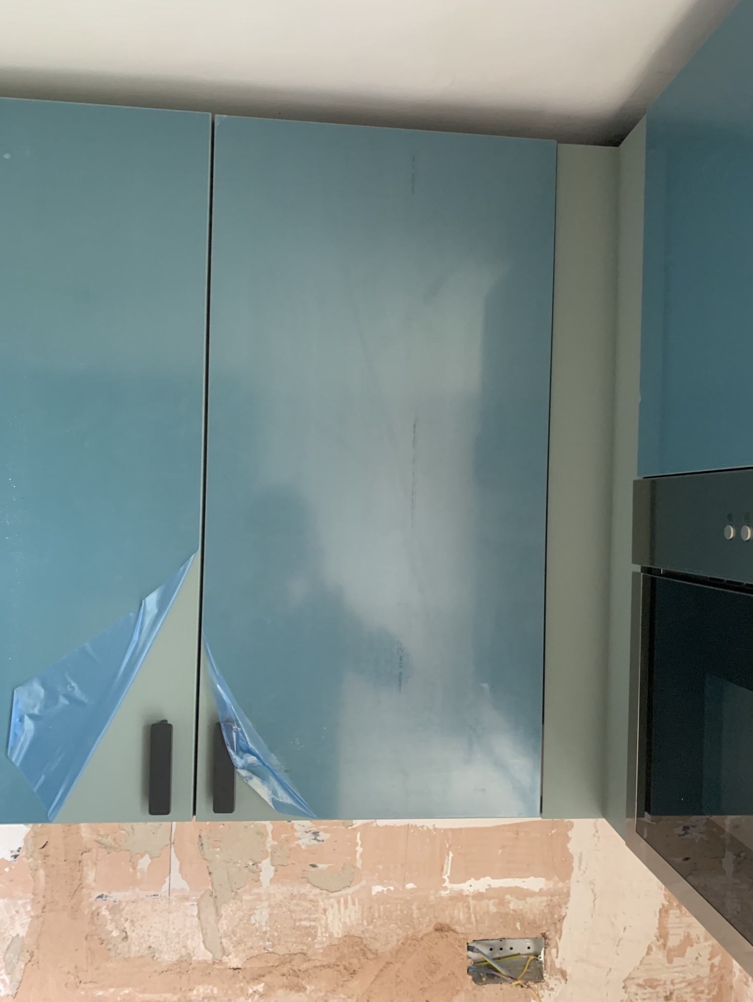 Niebieskie szafki kuchenne z folią ochronną częściowo odklejoną, widoczne ślady po remoncie na ścianie poniżej.