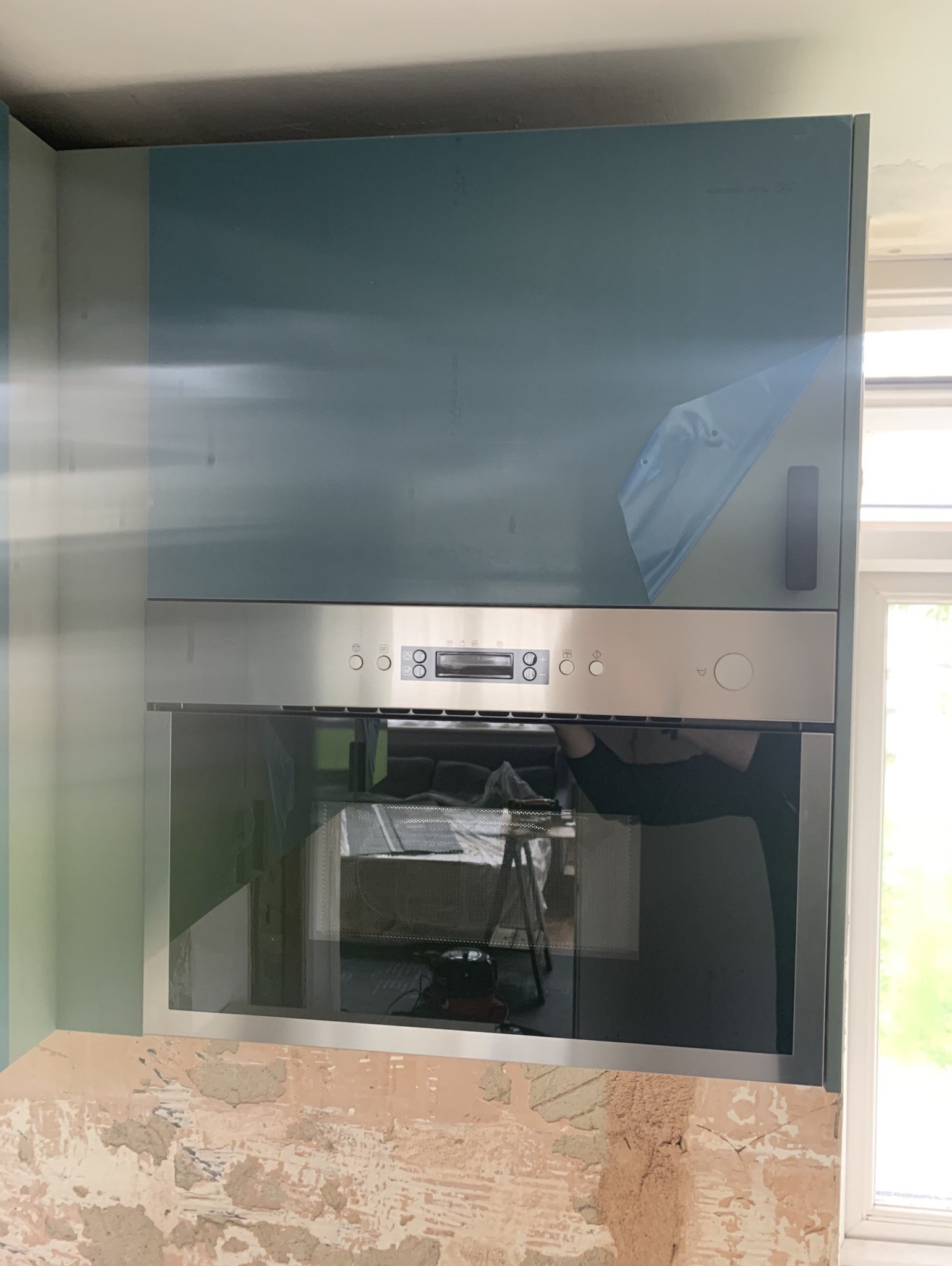 Nowa zabudowa kuchenna z piekarnikiem w trakcie montażu, widoczne ślady po szpachlowaniu na ścianie i folia ochronna na szafce.