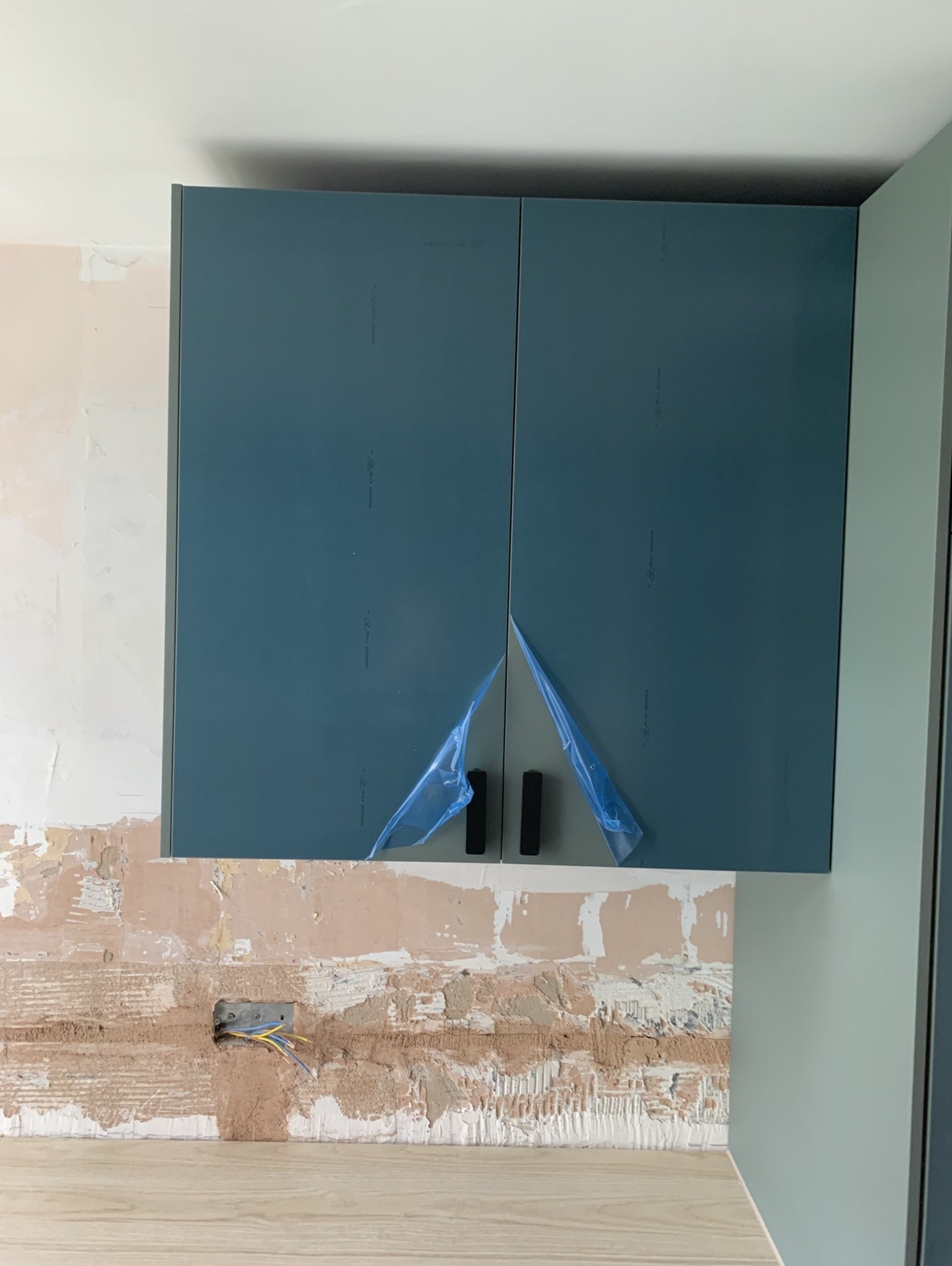Niebieskie szafki kuchenne z czarnymi uchwytami, częściowo zabezpieczone folią ochronną, zamontowane na ścianie z widocznymi śladami po remoncie i wystającymi przewodami elektrycznymi.