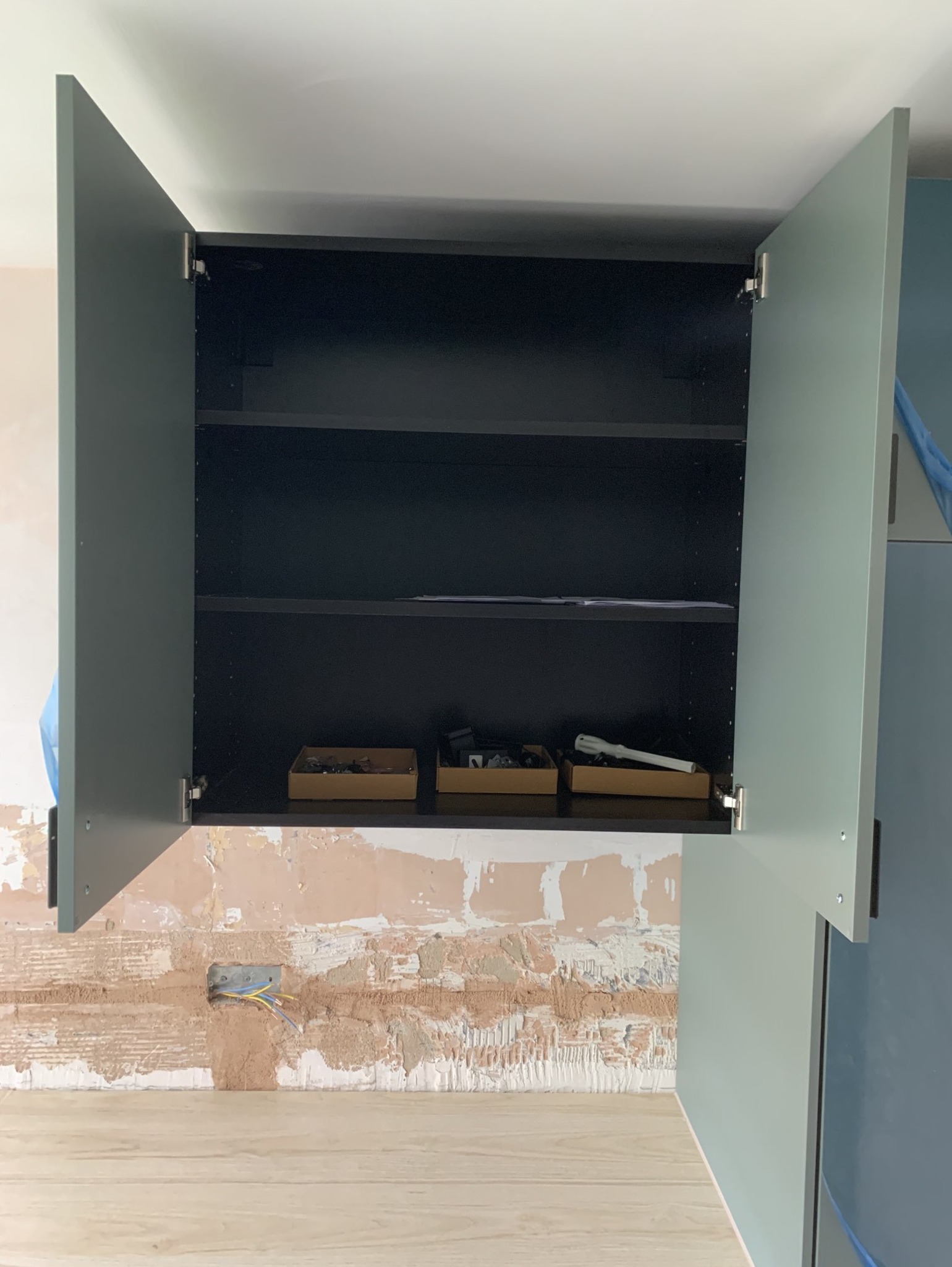 Montaż szafki kuchennej z otwartymi drzwiczkami, widoczne wnętrze z półkami i narzędziami w pudełkach, ściana w trakcie remontu z odsłoniętą instalacją elektryczną.