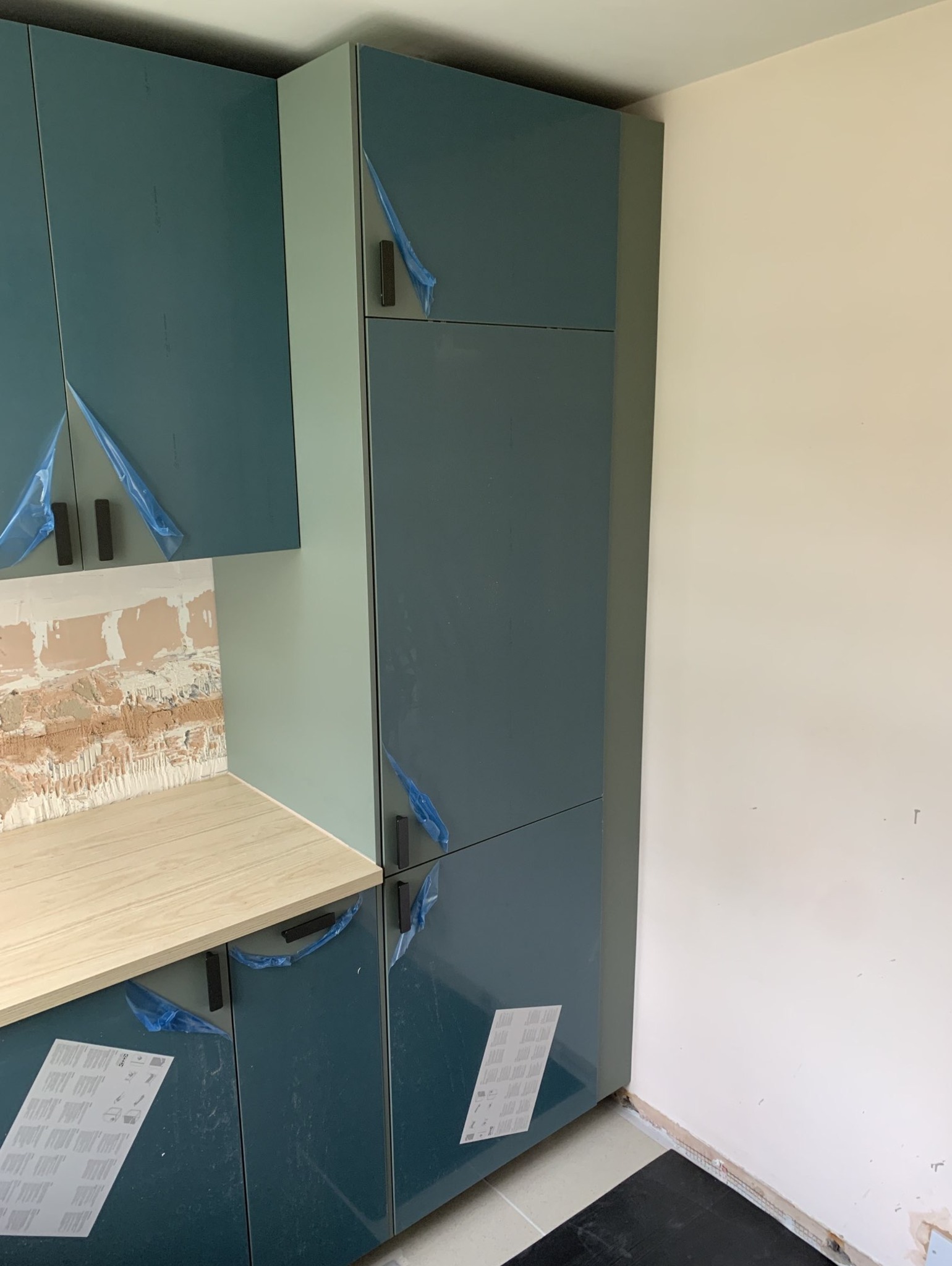 Nowe, granatowo-zielone szafki kuchenne z drewnianym blatem w trakcie montażu w Katowicach, zabezpieczone folią ochronną, z widoczną surową ścianą nad blatem.