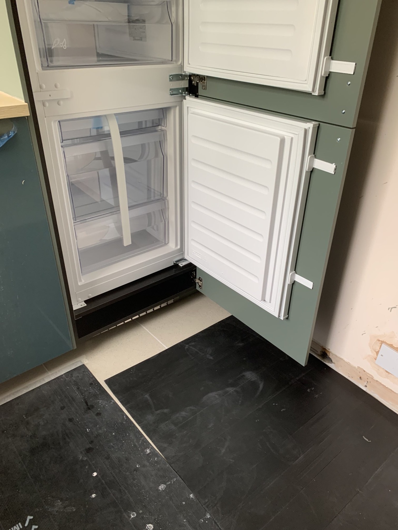 Wbudowana lodówka z otwartymi drzwiami, widoczne półki i szuflady, zamontowana w szafce z frontami w kolorze zielonym, czarna podłoga zabezpieczona kartonem.