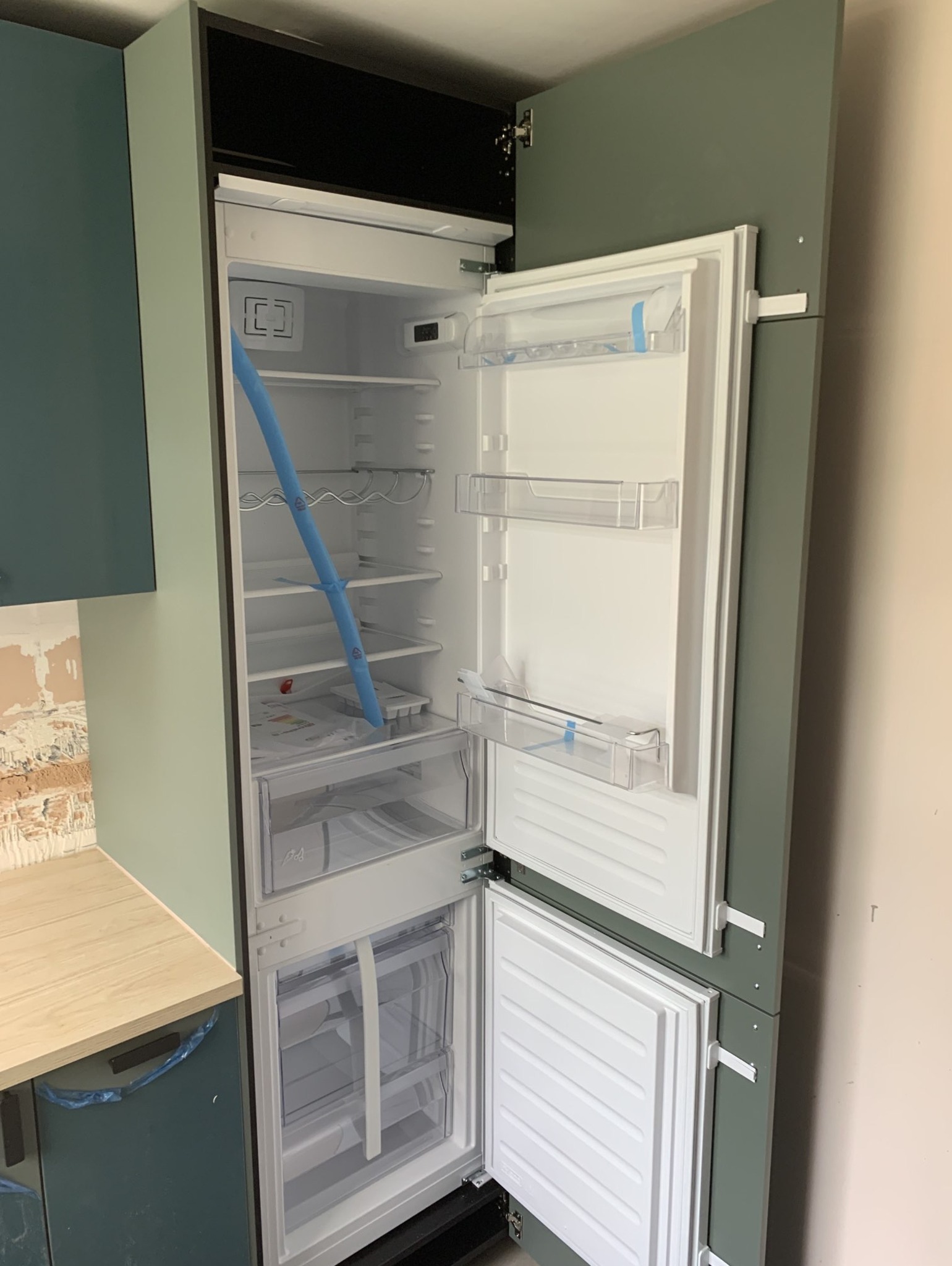 Wbudowana lodówka z otwartymi drzwiami, widoczne zabezpieczenia transportowe wewnątrz, zabudowa meblowa w odcieniach zieleni, fragment ściany z ceglastym wzorem.