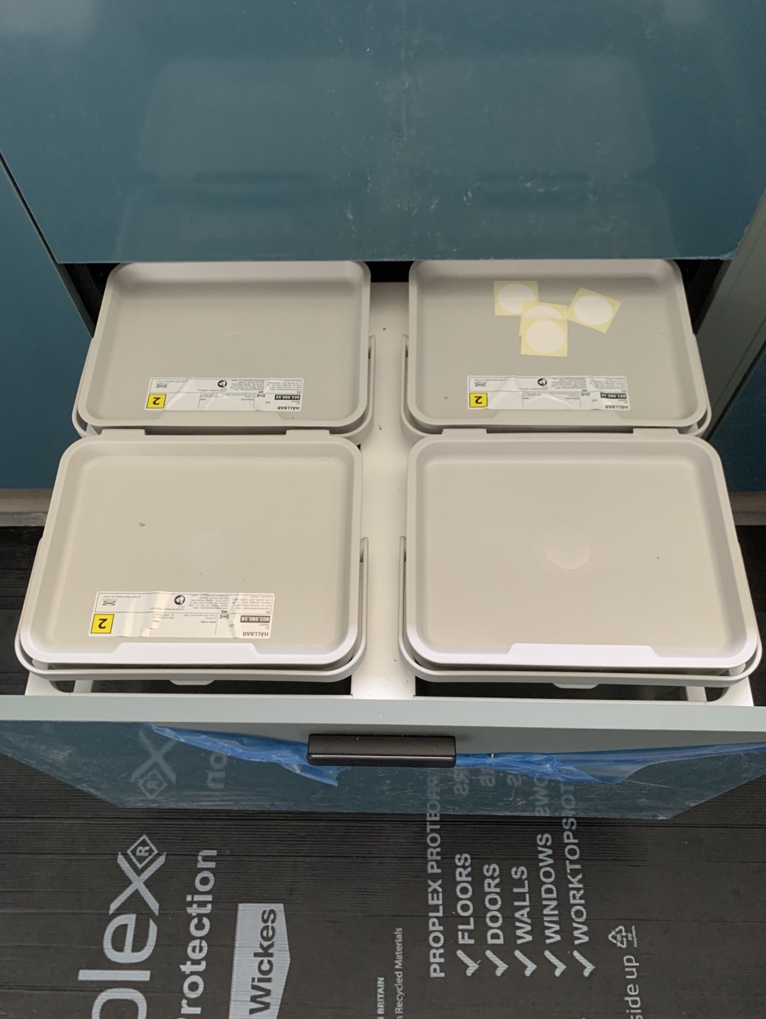 Otwarta szuflada szafki z czterema pojemnikami z tworzywa sztucznego, częściowo oklejonymi naklejkami i etykietami, na tle czarnej folii ochronnej z nadrukiem dotyczącym ochrony podłóg, ścian i drzwi.