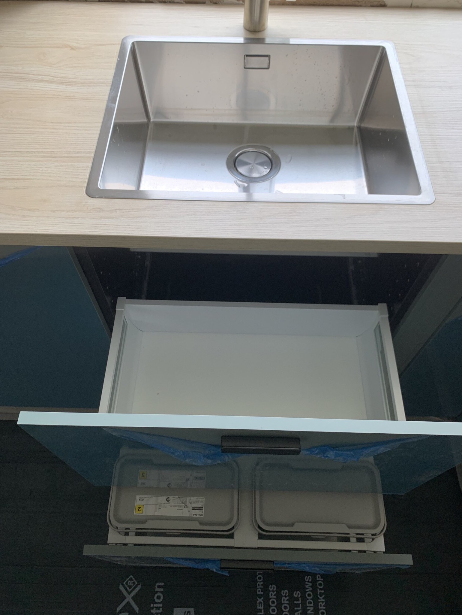 Zlewozmywak ze stali nierdzewnej w jasnym blacie, z wysuwaną szufladą pod spodem, zawierającą dwa pojemniki na odpady, zabezpieczone folią ochronną.